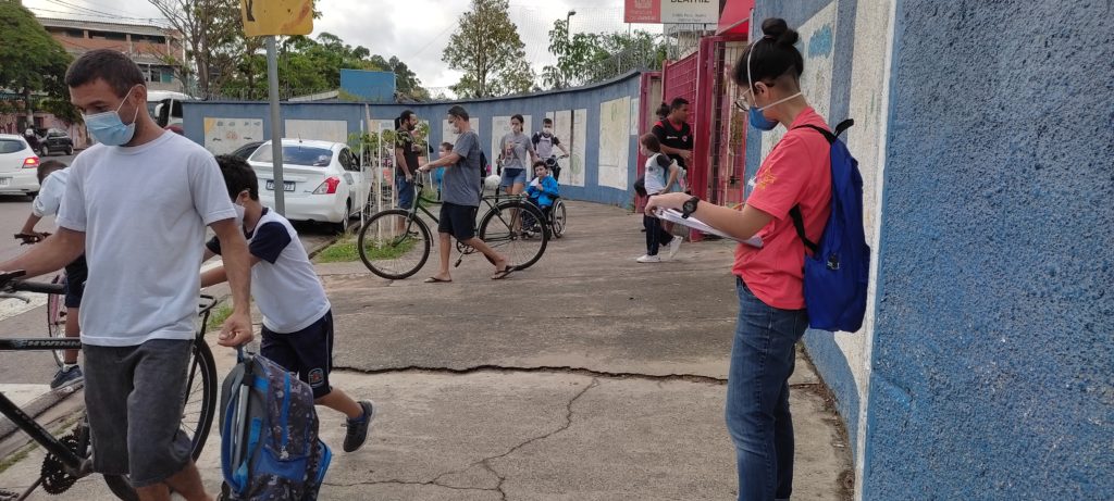 Grupo de pessoas em frente à saída de escola. À direita há um muro azul e branco com uma mulher de camiseta rosa encostada. À esquerda há um homem de camiseta branca e máscara de proteção facial empurrando uma bicicleta.
